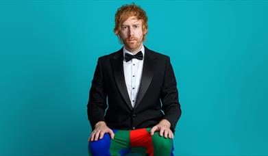 Mark Simmons sits in front of a blue backdrop in a black and white suit jacket but with colourful red blue and green medieval jester leggings and shoe
