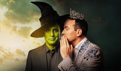 one man in pink jeweled blazer wearing a tiara, whispering in ear of a man dressed as Elphaba