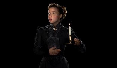 Woman in all black with blonde hair, holding candle, looking scared