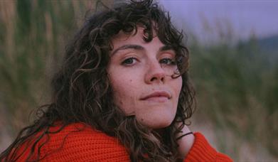 Ellie Gowers, in red knit jumper, with curly brown hair, slightly in face. Looking at camera with a slight smile