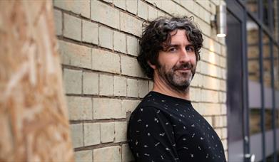 Mark Watson leaning against brick wall, black tshirt, smiling at camera