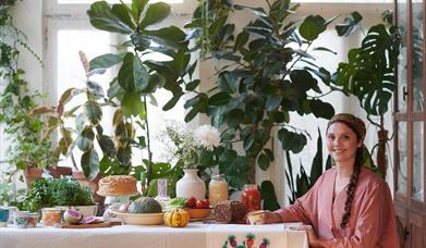 A young woman in pink with dark, plaited hair sits by a colourful display of fresh food and plants