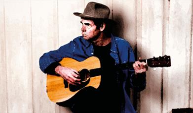 Rich Hall leaning against wall, playing acoustic guitar.