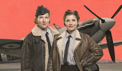 2 girls in pilot uniforms smiling at camera. Spitfire plane behind them