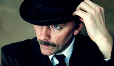 Man holding bowler hat on head, with a moustache, looking down at ground with a serious expression